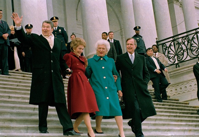 Barbara Bush memilih coat biru kehijauan dengan detail kancing besar untuk inaugurasi George H.W. Bush pada 1989. Busana kreasi desainer Bill Blass ini menunjukkan kesan klasik dan praktis dalam gaya busananya. Adapun Nancy Reagen ketika menyambut pasangan Bush tetap setia dengan warna merahnya.  (Foto: AP Photo/J. Scott Applewhite)