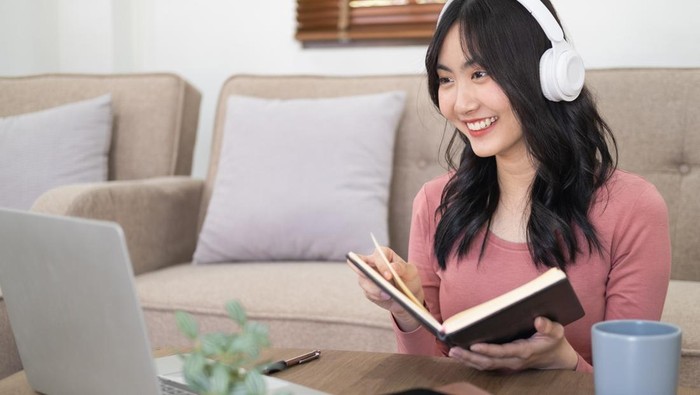 Smiling girl student wear wireless headphone study online with skype teacher, happy young woman learn language listen lecture watch webinar write notes look at laptop sit in cafe, distance education