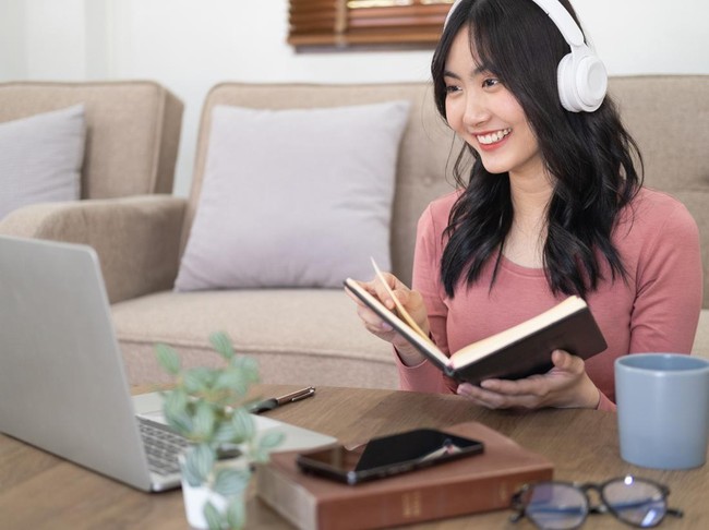 Smiling girl student wear wireless headphone study online with skype teacher, happy young woman learn language listen lecture watch webinar write notes look at laptop sit in cafe, distance education
