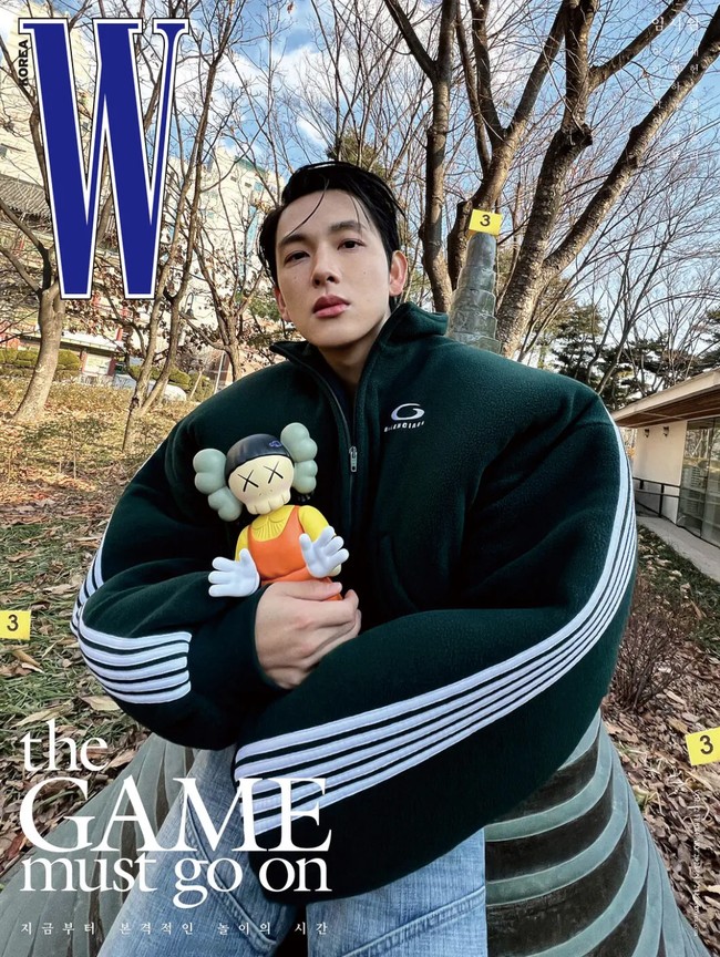 Ada pula penampilan Im Siwan memakai round neck zip-up jacket dengan detail puff pada bagian bahu. Warna jaketnya serasi dengan seragam peserta permainan bertahan hidup Squid Game. Tampak vinyl figure Younghee keluaran Kaws bikin salfok. Foto: dok. W Korea