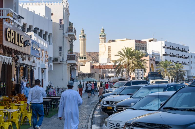 Muscat, Oman - 2 January 2024: A lively street scene captures locals and visitors amidst traditional architecture, with the iconic mosque towers standing tall against the clear blue sky.