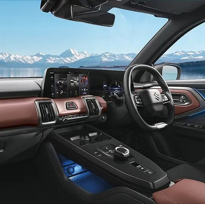 Sebuah SUV listrik Suzuki e-Vitara berwarna putih metalik diparkir di jalan perkotaan yang modern, dengan latar belakang gedung-gedung pencakar langit dan pepohonan hijau. Lampu depan LED-nya menyala terang, menyoroti desain futuristiknya.