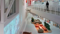 Ini penampakan koridor kantor yang serba putih, tentunya dengan aksen warna oranye. Terlihat ada beragam bentuk dan ukuran sofa di tengah ruangan.