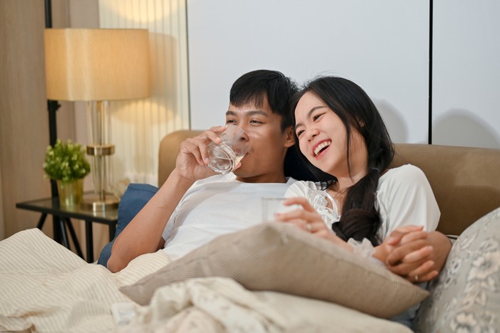 Ilustrasi suami istri A happy young Asian couple in pajamas is enjoying watching television or a movie together in bed. couple and domestic life concepts