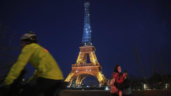 3 Tahun Perang Ukraina, Menara Eiffel Dihiasi Cahaya Biru-Kuning Ukrainians hold Ukrainian and European flag as the Eiffel Tower is illuminated with the colors of Ukraine to mark the third anniversary of Russia's invasion of the country, in Paris, Monday, Feb. 24, 2025. (AP Photo/Christophe Ena)