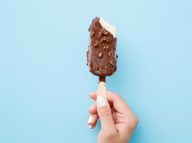 Young woman hand holding white vanilla ice cream with nuts and chocolate glaze on pastel light blue background. Bitten food. Closeup. Point of view shot. Top view.
