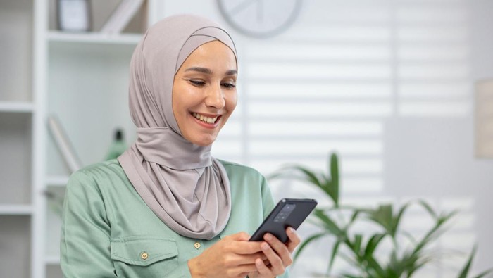 Cheerful Muslim woman wearing a hijab engages with her smartphone, portraying connectivity and modern lifestyle.