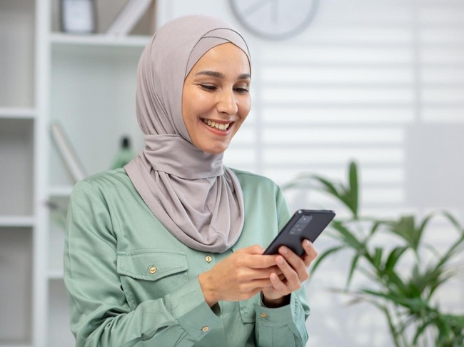 Cheerful Muslim woman wearing a hijab engages with her smartphone, portraying connectivity and modern lifestyle.