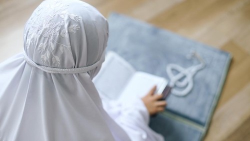 Rear View Of Young Muslim Girl Reading Quran On Prayer Mat