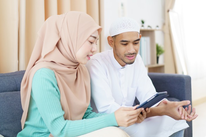Asian Muslim couple is sitting together in the living room, reading and studying the Quran during the fasting month of Ramadan. Asian muslim man and woman are reading the Koran on a digital tablet.