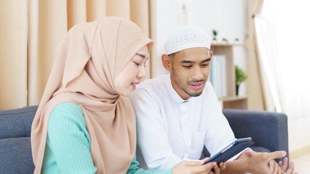 Asian Muslim couple is sitting together in the living room, reading and studying the Quran during the fasting month of Ramadan. Asian muslim man and woman are reading the Koran on a digital tablet.
