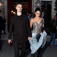 Romeo Beckham tampil monokrom dengan jas dan celana hitam, dipadu dalaman berwarna senada. Dia tampak membawa clutch merah marun sambil bergandengan tangan dengan Kim. Foto: GC Images/Pierre Suu