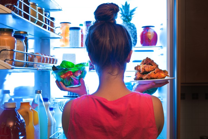 A young woman standing in front of the refrigerator, holding a bowl of fresh vegetable salad for a healthy diet on one hand and holding a plate of unhealthy fried chicken on the other. Making decision and choices for lifestyle and eating habit.