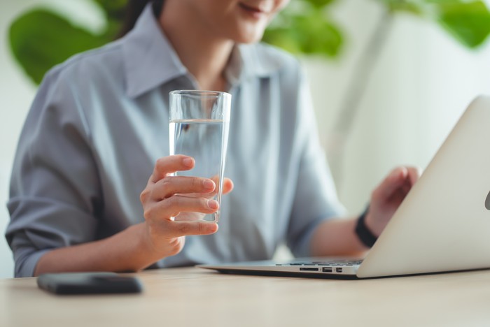 Ilustrasi minum air putih Close-up shot of woman relaxing taking a break drinking water while using laptop sitting at home office.