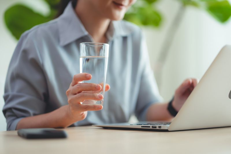 Close-up shot of woman relaxing taking a break drinking water while using laptop sitting at home office.