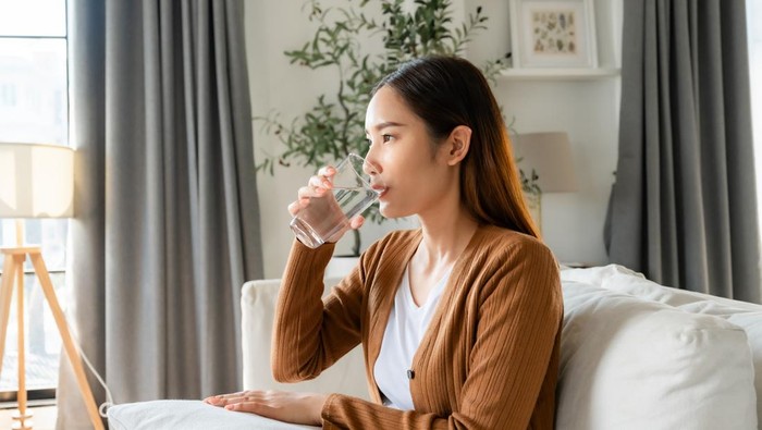 Happy young Asian woman drinking water while sitting on the sofa in the living room