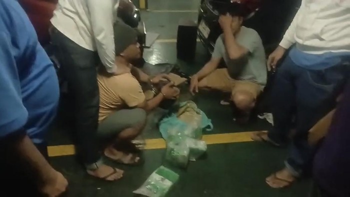 Tersangka Niko berpeci, berbaju coklat dan diborgol.