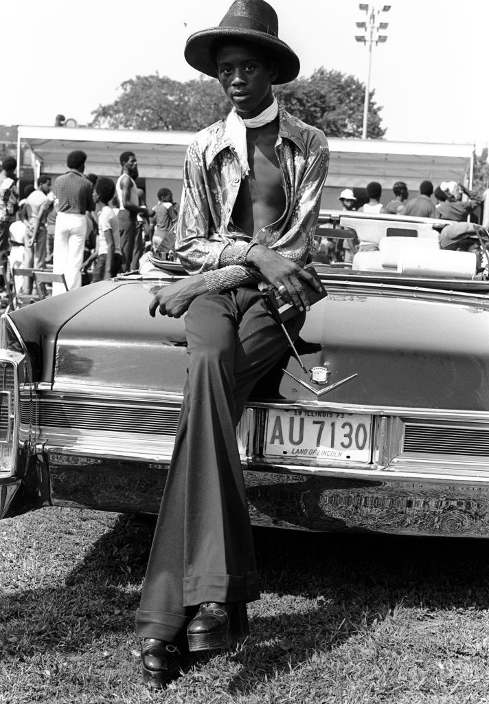 A young fashionably dressed African American man leans on the trunk of a Cadillac convertible, while enjoying a day outdoors in a Chicago park, 1973. He is wearing a hat, light scarf, an unbuttoned printed shirt, flare trousers, and platform shoes. (Photo by Robert Natkin/Getty Images)