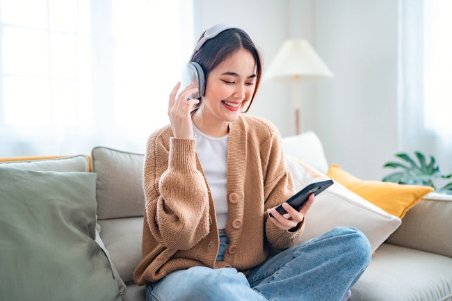 Ilustrasi wanita mendengar musik Happy young asian woman relaxing and listening music and watches movie on mobile phone with headphones while sitting on a couch in the morning at home