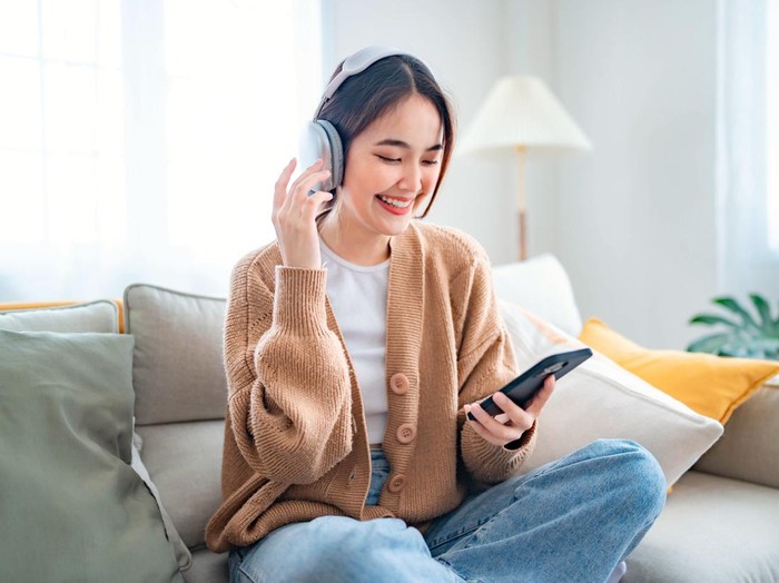Happy young asian woman relaxing and listening music and watches movie on mobile phone with headphones while sitting on a couch in the morning at home