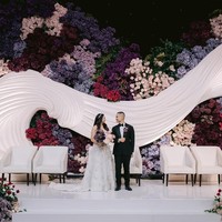 Pernikahan Cellos dan Natasha Putri terlihat mewah dengan dekorasi modern elegan ditambah sentuhan floral dramatis. Backdrop utama pelaminan Cellos menampilkan bentuk struktur putih bergelombang yang memberikan kesan dinamis dan mewah.  Foto: Dok. Instagram by.hilda