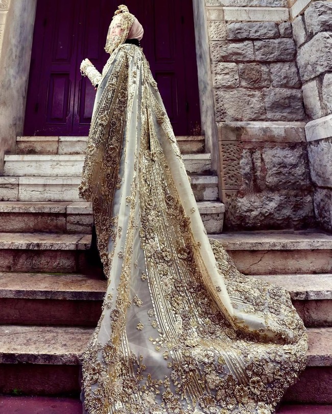 Outer berupa jubah panjang menjuntai hingga menyentuh lantai, dihiasi sulaman emas dan aplikasi tiga dimensi, membentuk motif floral dan garis vertikal yang memanjangkan siluet tubuh. Foto: Instagram/@princessyahrini @aldiphoto