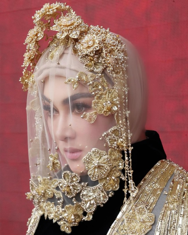 Penutup kepala didesain dengan veil transparan berhias aplikasi bordir bunga emas dan untaian mutiara kecil. Hiasan menyerupai mahkota floral berkilau yang dibentuk dengan teknik metalwork, menghadirkan nuansa fantasi dan regalia. Foto: Instagram/@princessyahrini @aldiphoto