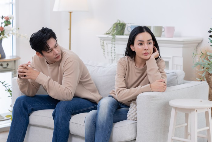 A couple sits on a couch  looking upset. One partner covers their face with their hands, while the other looks away, displaying clear signs of tension and disagreement