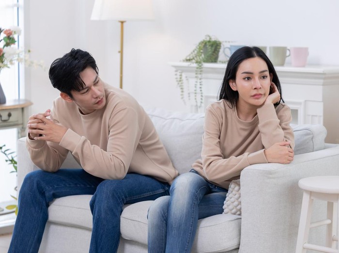 Ilustrasi pasangan suami istri A couple sits on a couch looking upset. One partner covers their face with their hands, while the other looks away, displaying clear signs of tension and disagreement