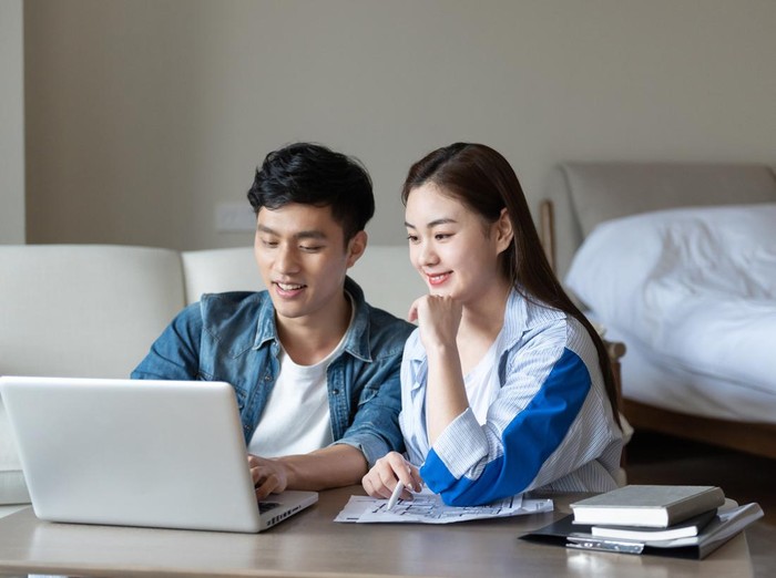 A young asian couple is sitting on a carpet using a computer