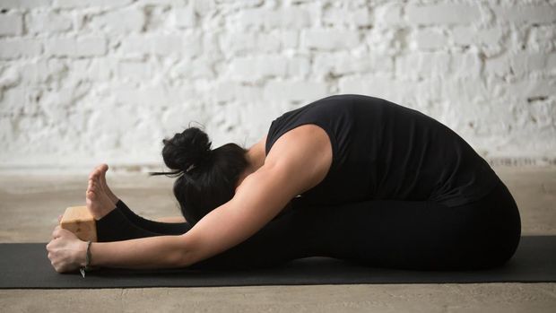 Young yogi woman practicing yoga concept, sitting in paschimottanasana exercise, using wooden block, seated forward bend pose, wearing black sportswear, working out, full length, white loft background