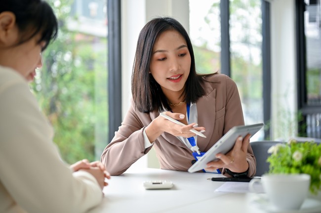 Ilustrasi wanita bekerja di kantor Beautiful and focused millennial Asian businesswoman using tablet, discussing work and working with her colleague in the office.