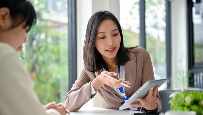 Beautiful and focused millennial Asian businesswoman using tablet, discussing work and working with her colleague in the office.
