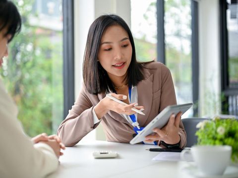 Beautiful and focused millennial Asian businesswoman using tablet, discussing work and working with her colleague in the office.