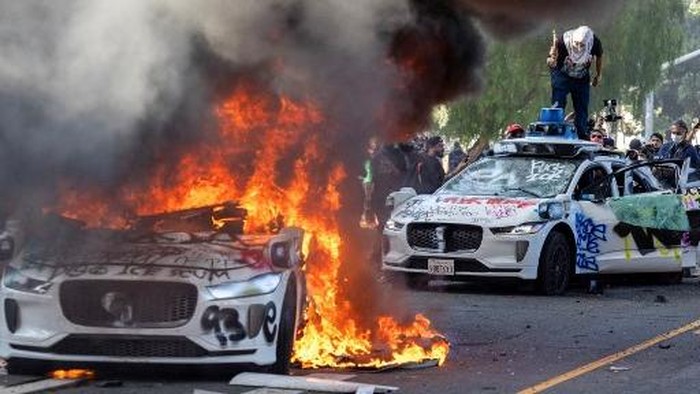  People drive past a burning Waymo car during immigration raid protests June 08, 2025 in Los Angeles, California. Tensions in the city remain high after the Trump administration called in the National Guard against the wishes of city leaders following two days of clashes with police during a series of immigration raids.   Mario Tama/Getty Images/AFP (Photo by MARIO TAMA / GETTY IMAGES NORTH AMERICA / Getty Images via AFP)