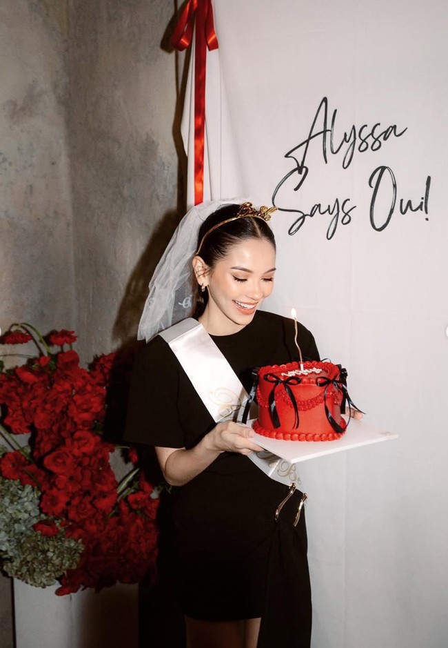 Tak seperti acara bridal shower yang menggunakan kue bergambar jorok, kue bridal shower Alyssa justru tampak simpel dan elegan dengan warna merah dan berhias pita-pita hitam. Dekorasi backdrop di acara tersebut juga tak kalah simpel dengan warna putih dan tulisan Alyssa Says Oui!. Foto: Instagram/@alyssadaguise