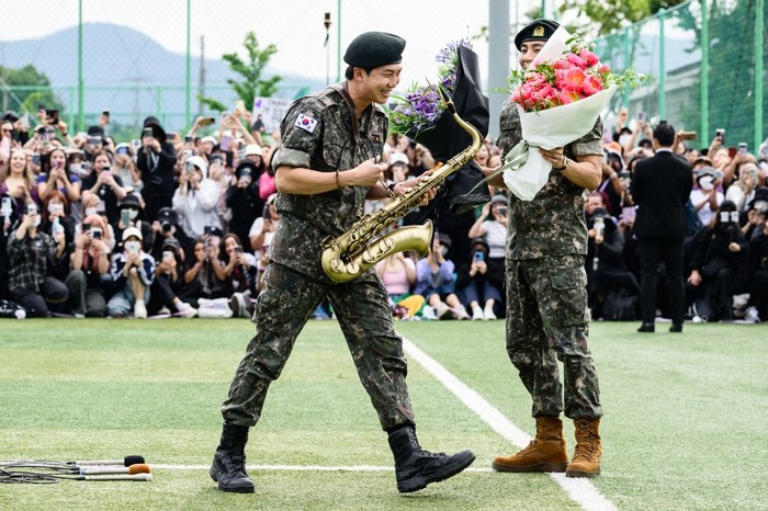 K-pop boy band BTS members RM (L) smiles while holding a saxophone while V looks on while holding flower bouquets at an outdoor sporting facility in Chuncheon on June 10, 2025, after their release from 18 months of South Korean military service. Hundreds of fans gathered near two army bases just outside of the capital Seoul where band leader RM -- recently named 