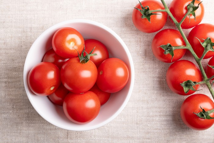 Red cherry tomatoes in a white bowl on linen fabric. Fresh, ripe type of small and round cocktail tomatoes, on the left side still on the vine, from above. Solanum lycopersicum var. cerasiforme. Photo