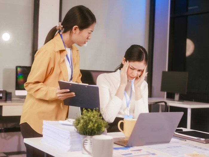 Two Asian women sit working at  desk,overwhelmed with stress struggling with headaches,feeling mentally and physically exhausted while trying to concentrate on their demanding tasks in a busy office