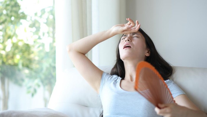 Stressed asian woman fanning suffering heat stroke sitting on a couch at home