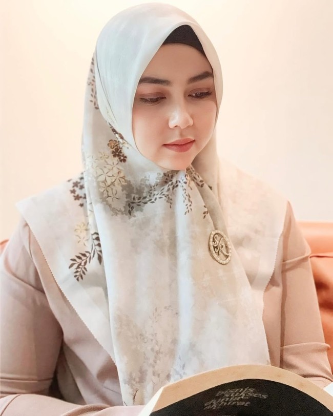Melalui akun Instagramnya, Lala Nurlela kerap mengenakan hijab segi empat warna pastel. Salah satunya saat sedang membaca buku ini, ia mengenakan hijab segi empat cream yang dibentuk menutupi bagian dada. Ia menambahkan bros dengan logo merek hijab Syahrini, SYR, di bagian dada. Foto: Dok. Instagram @lala_nurlelaa.