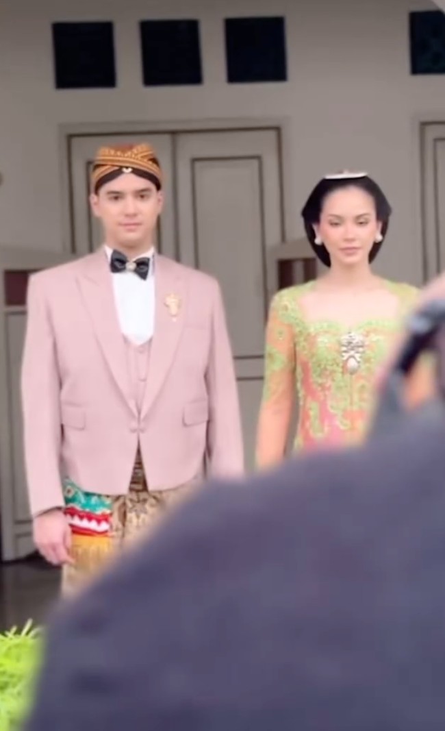 Di look kedua Alyssa dan Al tampil dengan busana adat Jawa modern yang lebih berwarna. Al tampak mengenakan jas yang dilengkapi dengan vest berwarna nude. Ia kemudian memadukannya dengan kemeja putih dan dasi kupu-kupu hitam. Sebagai bawahan, Al mengenakan kain lilit batik berwarna coklat. Foto: Instagram/@thebridedept