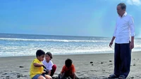 Terlihat dalam foto, Jokowi berdiri mengawasi tiga cucunya yang asyik bermain pasir pantai. Memakai kemeja putih dan celana hitam panjang, dia mengawasi cucuknya dengan seksama.  