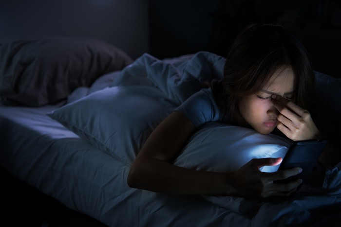 Young woman having sore and tired eyes when using a smartphone while lying in bed at night