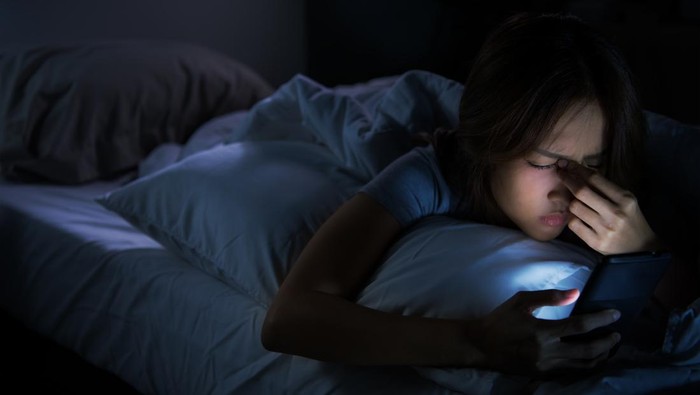 Young woman having sore and tired eyes when using a smartphone while lying in bed at night