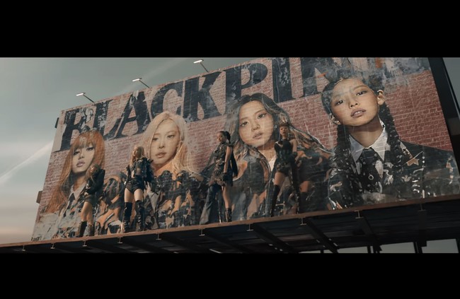 Video klip JUMP dibuka dengan adegan personel grup KPop BLACKPINK berkamuflase di depan billboard, tampil dalam balutan busana serba hitam berkonsep futuristik. Ada Jennie memakai corset rancangan The Delfos x Samuel Lewis, Lisa dengan puffer jacket Bent Kahina (Rp 72,8 juta), Jisoo memakai upcycled gloves dress Sevali, serta Rose dengan synthetic faux-leather jacket Noir Kei Ninomiya (Rp 80,4 juta). Foto: dok. YG Entertainment