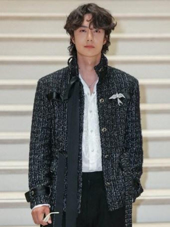 Wang Yibo mengenakan long tweed jacket hitam dari koleksi womenswear Chanel. Jaket tersebut memiliki detail tekstur woven khas maison Prancis, dengan sentuhan kilau halus yang mewah elegan. Foto: Thomas SAMSON / AFP