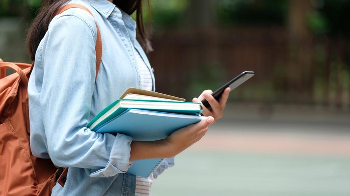 Student girl holding books and smartphone while walking in school campus background, education, back to school concept