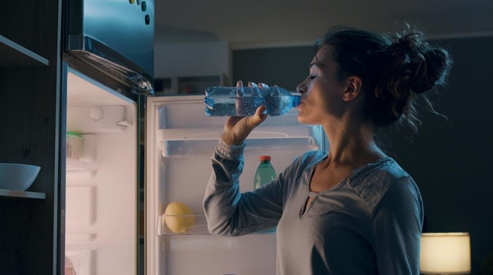 Thirsty woman standing in front of the fridge and drinking water late at night