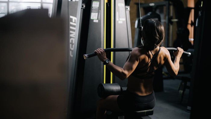 Muscular sporty woman using training simulator for pumping back muscles in gym, back of view. Concept of professional training in a modern dark gym.